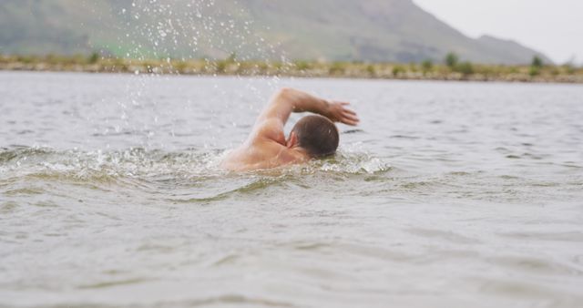 Man swimming in a natural lake with mountain scenery behind him. He is engaged in a swimming stroke, demonstrating endurance and physical fitness. Ideal for use in articles or advertisements related to outdoor activities, fitness, healthy lifestyles, and recreational pursuits.