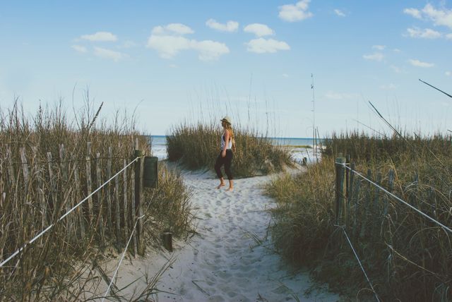 Depicts woman enjoying peaceful stroll on sandy beach path surrounded by tall grass. Ideal for promotional materials on travel, vacation getaways, nature retreats, calming and relaxation themes, and outdoor adventure content.