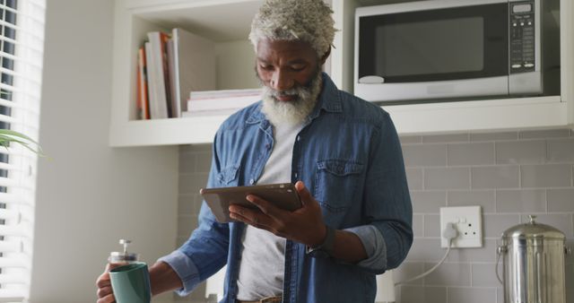 Mature man with grey hair and beard reading on a tablet while holding a mug in a modern kitchen. Ideal for lifestyle blogs, articles about senior engagement with technology, home appliance advertisements, and content depicting relaxed morning routines.