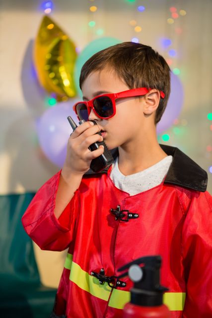 Boy dressed as fireman with red jacket and sunglasses, holding walkie-talkie at birthday party with balloons in background. Ideal for themes of childhood imagination, celebrations, and playful activities.