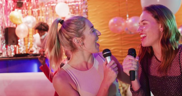 Two friends are singing karaoke at a party filled with balloons and festive decorations. Both look happy and engaged in singing using microphones, creating a lively atmosphere perfect for celebrations. Universal appeal for use in party invitations, social media posts, and lifestyle blogs emphasizing fun and enjoyment.