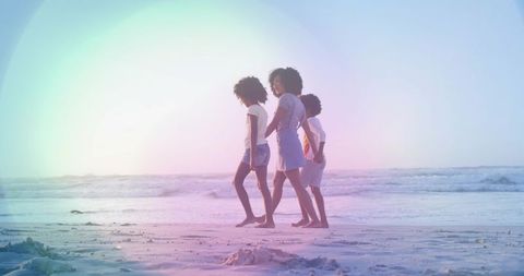 Walking mom leading two children along pastel beach shoreline at sunset with lens flare