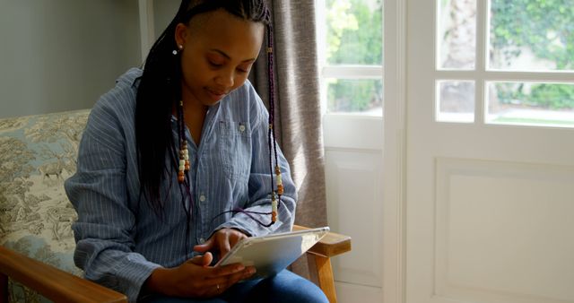 Young woman sitting on a wooden chair near a window, using a digital tablet. Ideal for themes related to technology, education, remote work, or modern home interiors. Can be used in blog posts, ads, or articles about digital devices, tech-savvy lifestyles, or online learning.