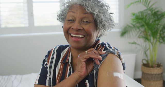 Senior woman proudly shows bandage on her arm after getting vaccinated, smiling joyfully in a well-lit room with plants. Useful for healthcare promotions, vaccination campaigns, senior wellbeing advertisements, medical articles, and self-care tips.