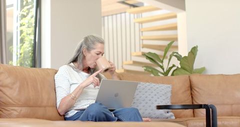 Mature Woman Enjoying Coffee While Browsing Laptop in Bright Living Room