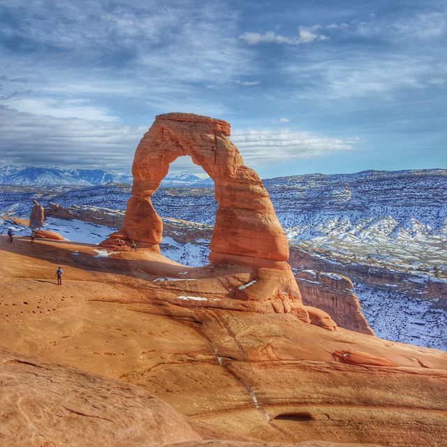 Hikers explore Delicate Arch, a famous natural sandstone arch in Arches National Park, Utah. The red rock formations and snow-covered ground highlight the contrast between the desert and winter elements. Useful for travel brochures, adventure blogs, and nature photography collections.