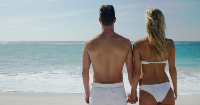 Couple wearing swimwear holding hands while standing on sandy beach looking at horizon. Ideal for vacation promotions, travel blogs, romantic getaways advertising, beachwear brands, summer-themed projects or social media content focused on relationships and travel.