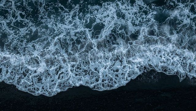 Aerial breaking waves moving across dark pebble shore with white foam filaments
