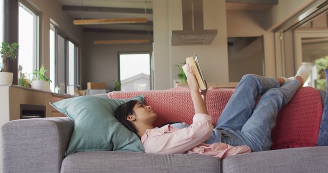 Image of biracial woman lying on sofa and reading book. Relax, leisure and spending free time at home with technology concept.