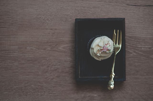 A beautifully decorated vanilla cupcake topped with creamy frosting and colorful sprinkles, presented with an elegant golden fork on a dark wooden tray. Ideal for use in food blogs, dessert recipes, culinary magazines, and social media posts showcasing artisanal baking.