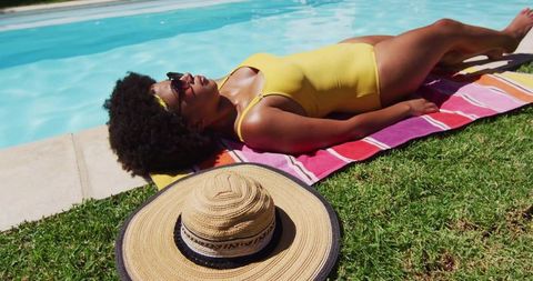 Sunbathing woman in yellow swimsuit beside turquoise pool on striped towel with straw hat