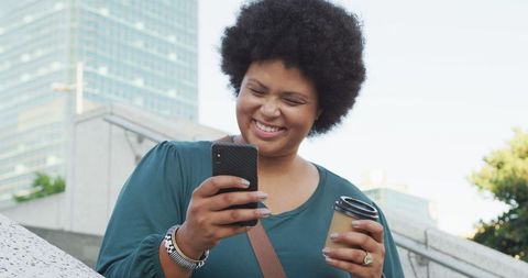 Smiling Woman Engaging with Smartphone Outdoors Holding Coffee