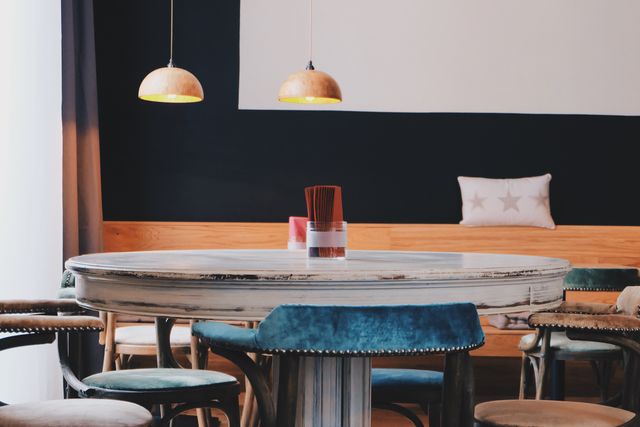 Modern café features round wooden table with chairs arranged around it. Soft pillow rests on bench seating against dark wall. Warm pendant lamps illuminate the scene with cozy ambiance. Ideal for articles on urban cafes, interior design inspiration, or hospitality industry trends.