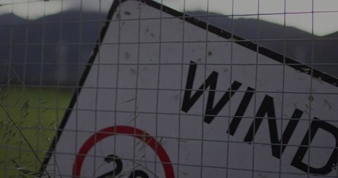 Leaning weathered wind warning sign behind wire mesh with tall grass in rural field