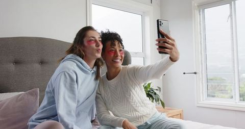 Diverse women taking selfie on bed wearing under-eye patches, cozy bedroom self-care moment