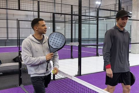 Diverse friends chatting on padel court holding racket and ball on purple indoor courts