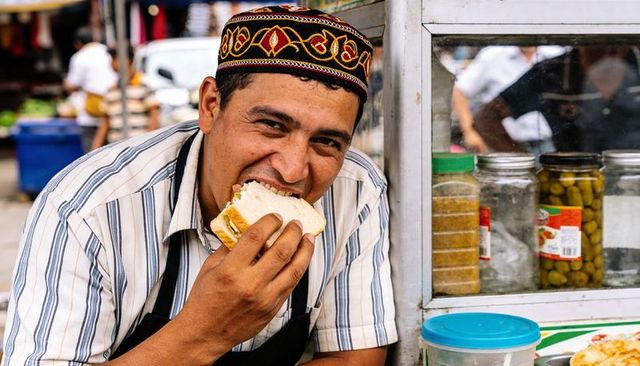 Smiling street vendor biting sandwich wearing embroidered cap at market stall with condiments