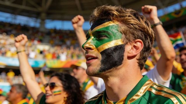 Male fan wearing green gold face paint cheering passionately in crowded stadium