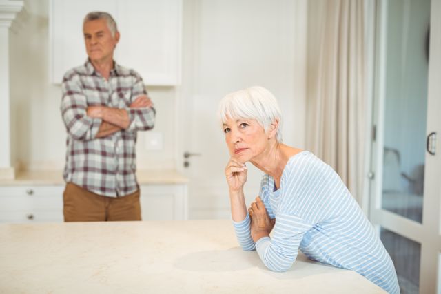 Elderly couple experiencing relationship issues in a modern kitchen. The woman is leaning on the counter, looking concerned, while the man stands in the background with arms crossed, indicating tension. Useful for articles on family dynamics, aging, relationship counseling, and emotional well-being.