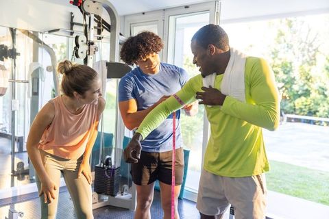Multiracial trainer measuring arm with tape while workout partners encourage in sunlit gym