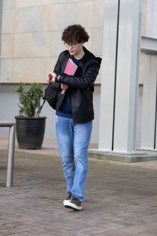 Teen male student walking across campus plaza checking watch holding pink folder