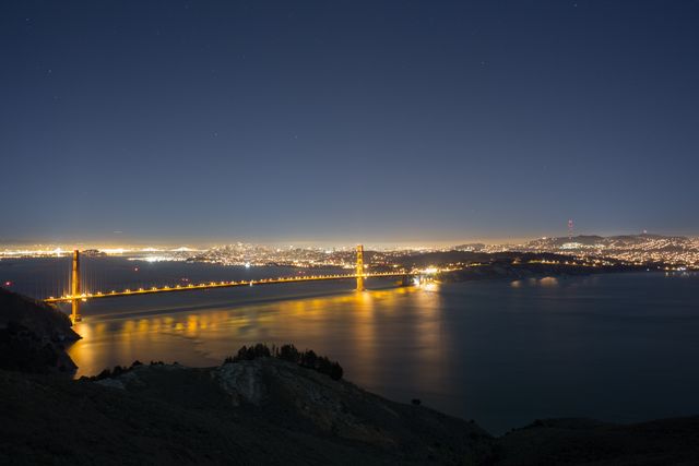 Golden Gate Bridge illuminated against a starry night sky with the brightly lit city skyline in the background. Tranquil bay waters reflecting the lights create a serene ambiance, perfect for use in travel brochures, architectural showcases, or scenic postcards.