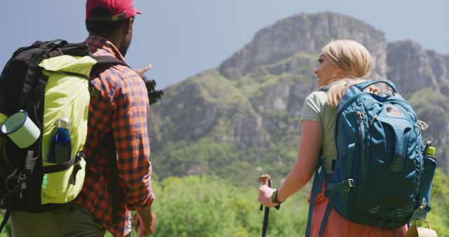 Diverse couple hiking on scenic mountain trail with lush green landscape, large backpacks, and trekking poles. Perfect for themes of adventure, travel, outdoor activities, diversity in nature, healthy lifestyle, and exploring majestic landscapes.