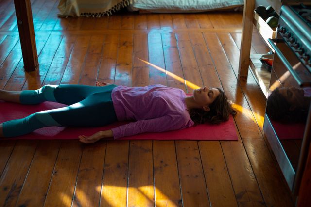 Woman lying on yoga mat in sunny cottage living room, eyes closed, appearing relaxed and peaceful. Ideal for promoting mindfulness, healthy living, and relaxation techniques. Suitable for wellness blogs, meditation guides, and lifestyle magazines.