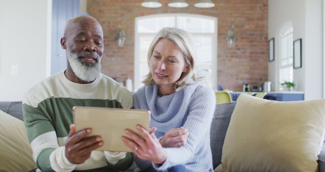 Happy senior diverse couple in living room sitting on sofa, using tablet, making image call. retirement lifestyle, at home with technology.