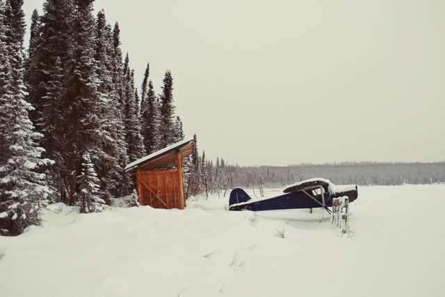 Snowy winter cabin next to an abandoned airplane, located in remote forest area. Ideal for travel blogs, nature documentaries, winter wilderness adventures, or promoting winter tourism. Evokes solitude, serenity, and the beauty of nature in cold environments.
