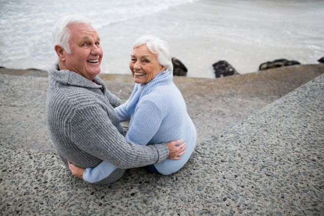 Senior couple sitting on a rocky beach, smiling and embracing each other. Ideal for use in advertisements or articles related to retirement, senior living, love and relationships, outdoor activities, and healthy aging.