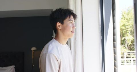 Asian man gazing out bedroom window morning light minimalist interior quiet contemplation