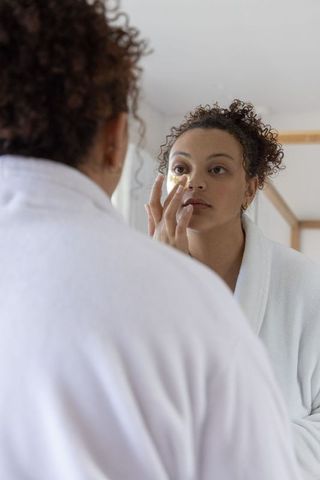African american woman applying gold under-eye patch in mirror during skincare routine