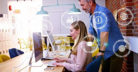 Woman and Mentor Collaborating at Desktop in Modern Open-Plan Office with Digital Icons