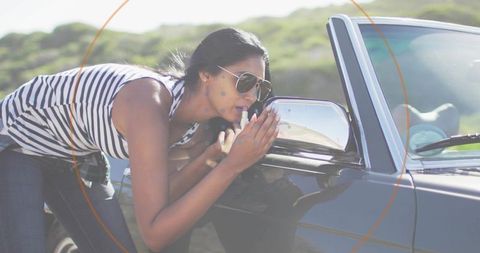 Woman leaning on convertible applying lipstick using side mirror during sunny roadside road trip