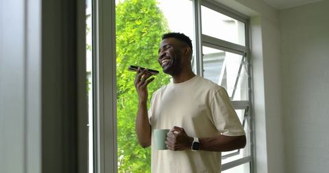 Smiling man speaking into smartphone while holding mug by bright window with green foliage