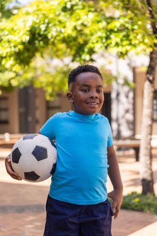 African american boy holding soccer ball in sunny courtyard smiling and ready to play
