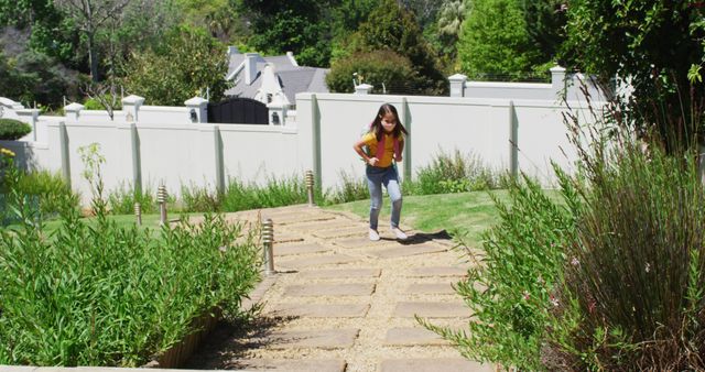 Young girl is walking along a pathway in a lush garden setting during a sunny day. The image includes green plants, shrubs, and a background with a clear sky, making it ideal for concepts related to outdoor activities, nature, childhood, and leisure. This image could be used in gardening magazines, children’s lifestyle blogs, or promotional material for outdoor recreation.