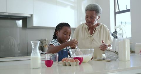 Grandmother guiding child whisking batter on bright modern kitchen island, baking together