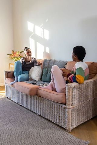 Two friends chatting and sipping coffee on cozy rattan sofa in sunlit living room