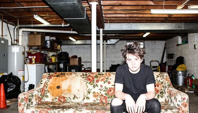 Disaffected young man sitting on stained floral sofa in messy basement with moody stare
