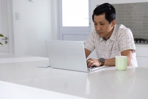 Asian man working on laptop at bright kitchen counter with mug, remote work
