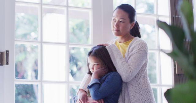 A concerned mother is comforting her daughter by the window. Both are standing indoors with natural light coming through the window. This scene depicts familial support, affection, and maternal care. Suitable for use in parenting articles, emotional support content, and family-related blogs.