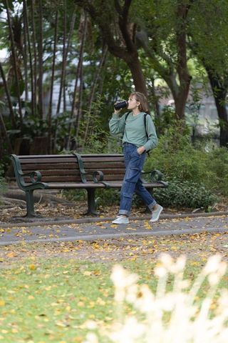 Young woman walking in autumn park sipping reusable bottle beside wooden bench