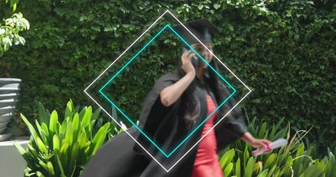 Graduate walking in garden wearing mortarboard and gown, talking on phone while holding diploma