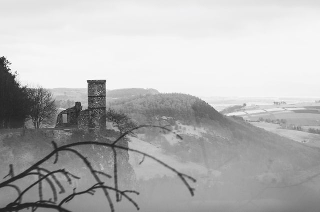 Photo captures a ruined castle tower perched on a hill overlooking a misty landscape, with distant hills visible in the background. The black and white toning adds a moody and atmospheric feel. Useful for projects related to history, travel, medieval architecture, and moody aesthetics.