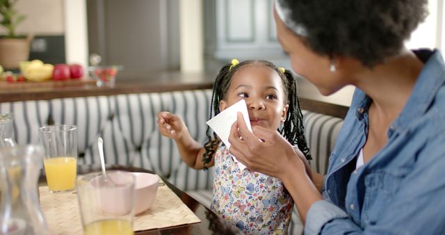 Mother gently cleaning her daughter's face while they have breakfast together. Use this to showcase family bonding moments, morning routines, or advertisements highlighting healthy, loving family environments.