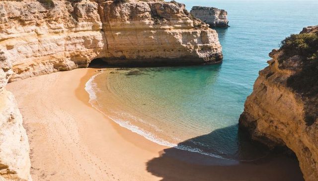 Sunlit limestone cliffs framing secluded golden beach and turquoise cove with sea cave