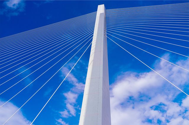 Image depicts a modern cable-stayed bridge tower with elegant cables extending against a clear blue sky with some clouds. Architectural and engineering excellence is highlighted in this image, showcasing structural design and urban development. It is suitable for use in articles, presentations, and educational materials focused on engineering, architecture, modern construction techniques, and infrastructure projects.