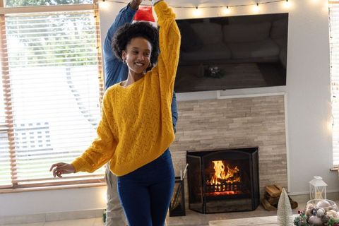 African american woman twirling in yellow sweater, partner holding santa hat by fireplace